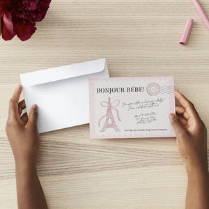 May include: A light pink baby shower invitation card featuring the text Bonjour B&eacute;b&eacute;, a pink Eiffel Tower illustration, and a postage stamp graphic. The card is paired with a plain white envelope, showcasing a scalloped edge design.