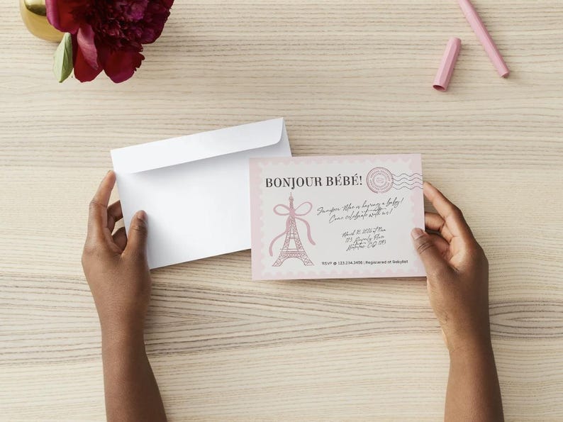 May include: A light pink baby shower invitation card featuring the text Bonjour B&eacute;b&eacute;, a pink Eiffel Tower illustration, and a postage stamp graphic. The card is paired with a plain white envelope, showcasing a scalloped edge design.