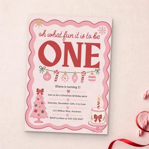 May include: A rectangular birthday invitation card with a scalloped pink border. The text reads: oh what fun it is to be ONE, Eliana is turning 1!, Join us for a Christmas birthday party, Saturday, December 12th, 2 to 4 pm, 111 Willow Way, Anytown, RSVP to Mom at 555-123-4567. Features include a pink Christmas tree, a cake with a candle, and a string of ornaments.