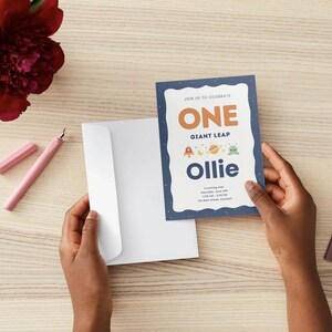 K&ouml;nnte beinhalten: Eine rechteckige Geburtstagseinladungskarte mit dunkelblauem Hintergrund und wei&szlig;em Wellenrand. Der Text lautet: JOIN US TO CELEBRATE ONE GIANT LEAP Ollie is turning one. Sie zeigt kleine weltraumbezogene Symbole wie eine Rakete und einen Planeten.