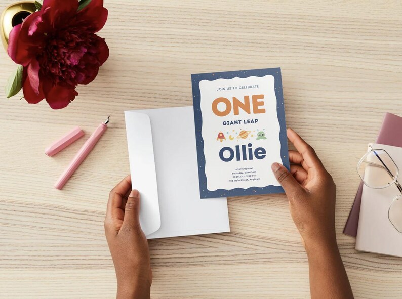 K&ouml;nnte beinhalten: Eine rechteckige Geburtstagseinladungskarte mit dunkelblauem Hintergrund und wei&szlig;em Wellenrand. Der Text lautet: JOIN US TO CELEBRATE ONE GIANT LEAP Ollie is turning one. Sie zeigt kleine weltraumbezogene Symbole wie eine Rakete und einen Planeten.