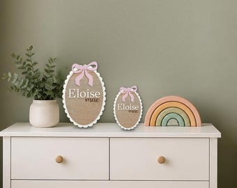 May include: Two oval wooden plaques with scalloped white edges and pink bows, each featuring the name Eloise Mae in white and brown lettering. The plaques are displayed alongside a small potted plant and a multi-colored arched rainbow.