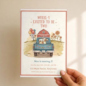 May include: A rectangular birthday invitation card featuring a blue pickup truck carrying a goat, balloons, and a red barn. The text reads: WHEEL-Y EXCITED TO BE TWO, Max is turning 2!, Saturday, June 14th, 11:00 AM - 2:00 PM, 123 Main Street, Anytown, RSVP to Mom @ 555.123.4567 by June 1st.