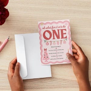 May include: A pink birthday invitation card with a scalloped edge featuring the text "oh what fun it is to be ONE" in red, decorated with illustrations of a Christmas tree, ornaments, and candy canes, paired with a plain white envelope.