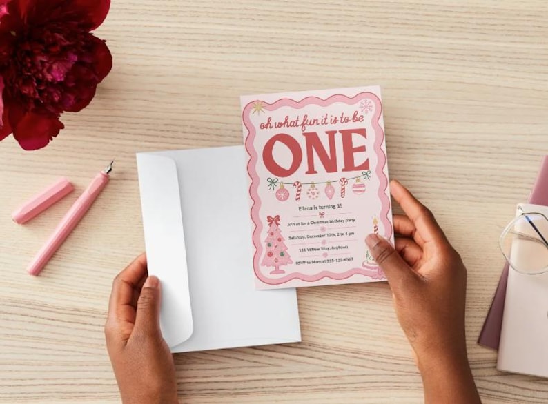 May include: A pink birthday invitation card with a scalloped edge featuring the text "oh what fun it is to be ONE" in red, decorated with illustrations of a Christmas tree, ornaments, and candy canes, paired with a plain white envelope.