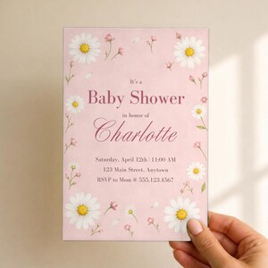 Puede incluir: Una tarjeta de invitaci&oacute;n rectangular para baby shower sobre un fondo rosa claro con ilustraciones de margaritas blancas y peque&ntilde;as flores rosas. El texto dice: It's a Baby Shower in honor of Charlotte, Saturday, April 12th, 11:00 AM, 123 Main Street, Anytown, RSVP to Mom @ 555.123.4567.
