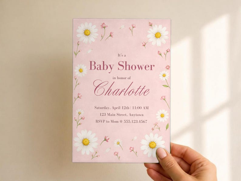 Puede incluir: Una tarjeta de invitaci&oacute;n rectangular para baby shower sobre un fondo rosa claro con ilustraciones de margaritas blancas y peque&ntilde;as flores rosas. El texto dice: It's a Baby Shower in honor of Charlotte, Saturday, April 12th, 11:00 AM, 123 Main Street, Anytown, RSVP to Mom @ 555.123.4567.