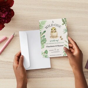 May include: A baby shower invitation card featuring a watercolor illustration of a baby dinosaur hatching from an egg, surrounded by green tropical leaves. The text reads A WILD ONE is on the way! Come celebrate the parents to be Charlotte & Mason AUGUST 7 7PM 123 ANYWHERE ST REGISTERED AT BABYLIST.