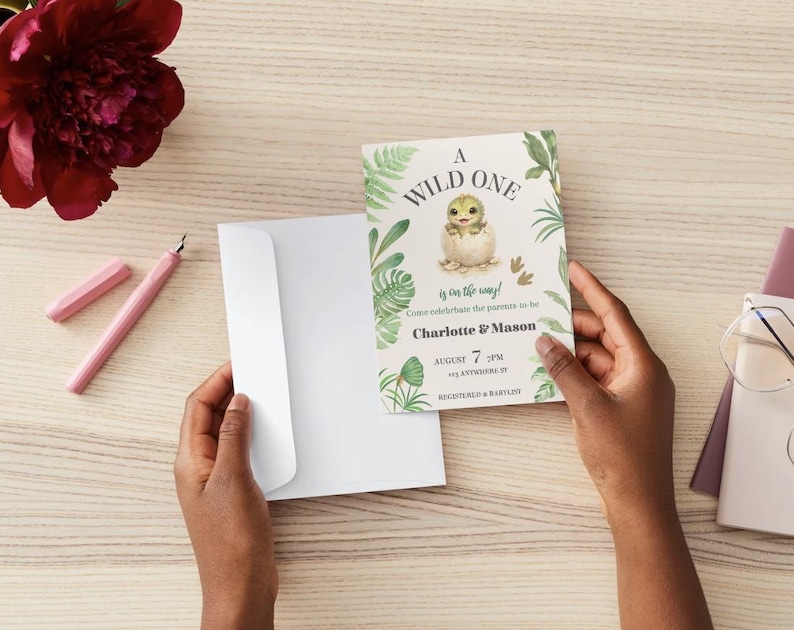 May include: A baby shower invitation card featuring a watercolor illustration of a baby dinosaur hatching from an egg, surrounded by green tropical leaves. The text reads A WILD ONE is on the way! Come celebrate the parents to be Charlotte & Mason AUGUST 7 7PM 123 ANYWHERE ST REGISTERED AT BABYLIST.