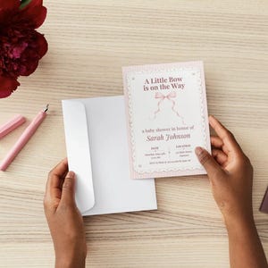 Puede incluir: Una tarjeta de invitaci&oacute;n rectangular para baby shower con un borde de rayas rosa claro y un gr&aacute;fico de lazo rosa en el centro. El texto dice A Little Bow is on the Way, a baby shower in honor of Sarah Johnson, con espacios para fecha y lugar.
