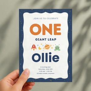 K&ouml;nnte beinhalten: Eine rechteckige Einladungskarte mit einem dunkelblauen, weltraumthematischen Rand mit Sternen. Die wei&szlig;e Mitte zeigt den Text JOIN US TO CELEBRATE, ONE GIANT LEAP, Ollie, is turning one, Saturday, June 14th, 11:00 AM - 2:00 PM, 123 Main Street, Anytown sowie Symbole einer Rakete, eines Mondes, eines Planeten und eines Au&szlig;erirdischen.