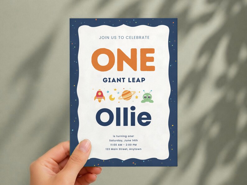 K&ouml;nnte beinhalten: Eine rechteckige Einladungskarte mit einem dunkelblauen, weltraumthematischen Rand mit Sternen. Die wei&szlig;e Mitte zeigt den Text JOIN US TO CELEBRATE, ONE GIANT LEAP, Ollie, is turning one, Saturday, June 14th, 11:00 AM - 2:00 PM, 123 Main Street, Anytown sowie Symbole einer Rakete, eines Mondes, eines Planeten und eines Au&szlig;erirdischen.