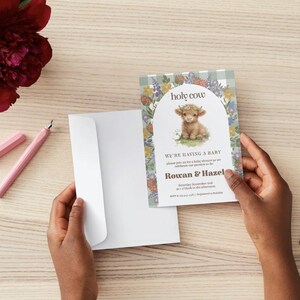 May include: A baby shower invitation card featuring a highland cow illustration, floral arch, and the text holy cow, we're having a baby, Rowan & Hazel. The card has a light green gingham border and is paired with a plain white envelope.