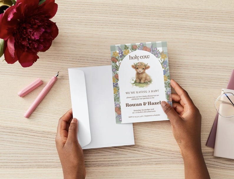 May include: A baby shower invitation card featuring a highland cow illustration, floral arch, and the text holy cow, we're having a baby, Rowan & Hazel. The card has a light green gingham border and is paired with a plain white envelope.