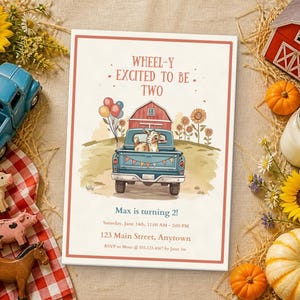 Könnte beinhalten: Eine rechteckige Geburtstagseinladungskarte mit einem blauen Pickup, der eine Ziege, eine rote Scheune und Sonnenblumen transportiert. Der Text lautet: WHEEL-Y EXCITED TO BE TWO, Max is turning 2!, Saturday, June 14th, 11:00 AM - 2:00 PM, 123 Main Street, Anytown.