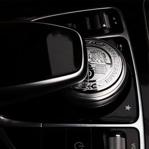 Puede incluir: Primer plano del panel de control interior de un coche. Un dial plateado con el texto "AFFALTERBACH" y un emblema de árbol está centrado. El panel es negro con detalles plateados y varios botones y diales, incluyendo "DYNAMIC" y un control de volumen.