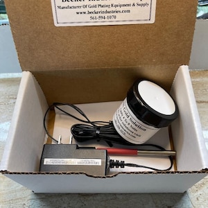 Puede incluir: Una caja blanca con un adaptador de corriente negro, un cable blanco y negro, un cable rojo y blanco y un pequeño frasco blanco con tapa negra. El frasco está etiquetado como "AK Gold Solution" y tiene el texto "Apply 4 to 8 Volts" y "Order solutions visit www.beckerindustries.com". La caja tiene una etiqueta en la parte superior que dice "Becker Industries Corp" y tiene el texto "Manufacturer of Gold Plating Equipment & Supply" y "www.beckerindustries.com" y "561-594-1070".
