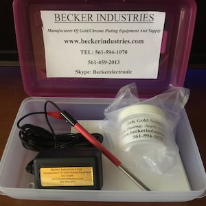 May include: A white plastic container with a pink lid, containing a small black power supply, a red and black wire, a silver pen, and a small white jar labelled "14K Gold Solution, Plating, Apply", with the text "Beckerindustries.com 561-594-1070" printed on the jar.