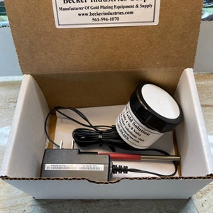 May include: A white box with a black power adapter, a red and silver probe, and a small jar labeled "AK Gold Solution". The box has a label on the top that reads "Becker Industries Corp. Manufacturer of Gold Plating Equipment & Supply www.beckerindustries.com 561-594-1070".