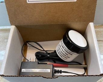 Op de afbeelding: Een witte doos met een zwarte adapter, een rode en zilveren sonde en een klein potje met het label "AK Gold Solution". De doos heeft een label aan de bovenkant met de tekst "Becker Industries Corp. Fabrikant van verguldingsapparatuur en benodigdheden www.beckerindustries.com 561-594-1070".