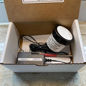 May include: A white box with a white interior containing a gold plating kit. The kit includes a small jar of gold solution, a power adapter, and a red and black wire. The box has a label on the top that reads "Becker Industries Corp. Manufacturer of Gold Plating Equipment & Supply www.beckerindustries.com 561-594-1070".