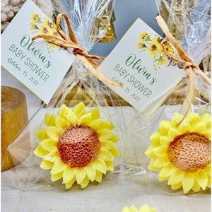 May include: Two small yellow and orange sunflower-shaped soaps are packaged in clear cellophane bags tied with twine. Each bag has a small white card with the text "Olivia's Baby Shower October 15, 2022".