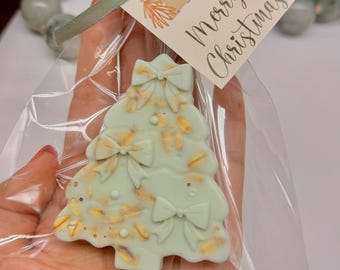 May include: A light green Christmas tree-shaped treat with bow details, packaged in clear cellophane and tied with a green ribbon. A tag with "Merry Christmas" is attached. The treat is held in a person's hand.