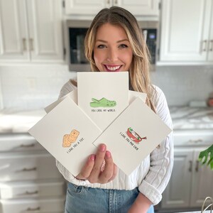 May include: Three greeting cards with illustrated puns. The first card shows a green and yellow clog with the text "You croc my world." The second card shows two orange tater tots with the text "I love you a tot." The third card shows a bowl of pho with chopsticks and the text "I love you pho ever."