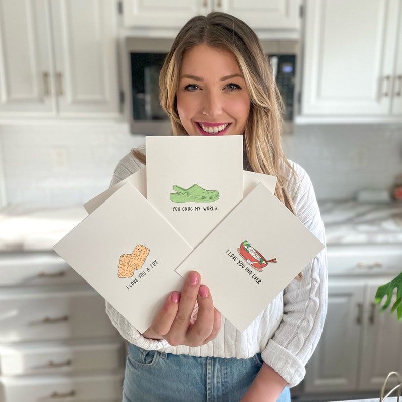 May include: Three greeting cards with illustrated puns. The first card shows a green and yellow clog with the text "You croc my world." The second card shows two orange tater tots with the text "I love you a tot." The third card shows a bowl of pho with chopsticks and the text "I love you pho ever."