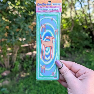 May include: Laminated bookmark featuring an alien dog design. The bookmark has a teal background with a swirling pattern and illustrations of the alien dog. The top of the bookmark has the text "One Durian" and "Alien Dog".