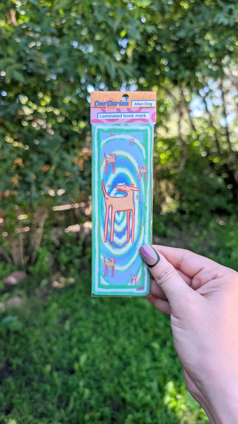 May include: Laminated bookmark featuring an alien dog design. The bookmark has a teal background with a swirling pattern and illustrations of the alien dog. The top of the bookmark has the text "One Durian" and "Alien Dog".