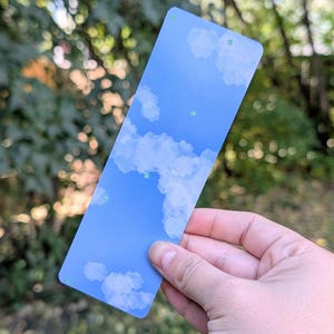 May include: A rectangular bookmark featuring a blue sky design with fluffy white clouds. The bookmark has rounded corners and small green dots. The background is blurred, showing green foliage.