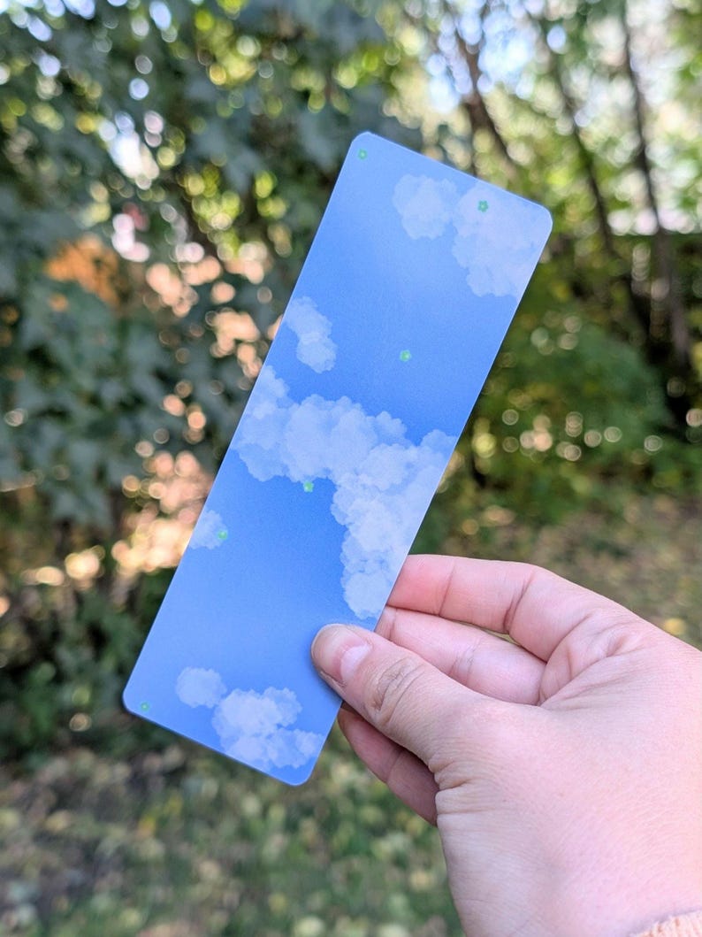 May include: A rectangular bookmark featuring a blue sky design with fluffy white clouds. The bookmark has rounded corners and small green dots. The background is blurred, showing green foliage.