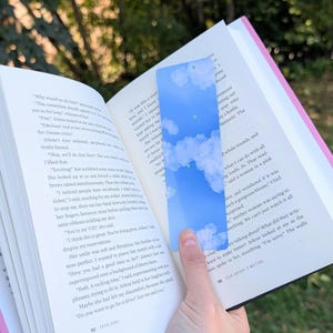 May include: A rectangular bookmark with a blue sky and cloud design. The bookmark is held open in a book with text visible. The bookmark is a light blue color with white clouds.