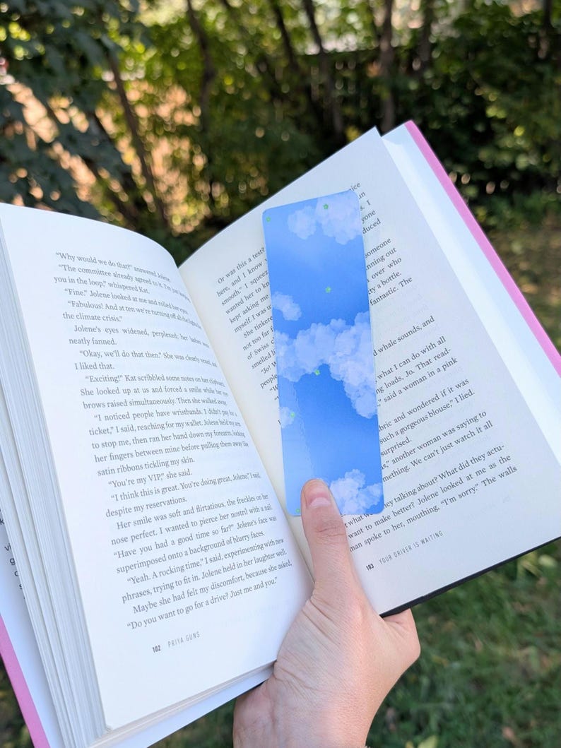 May include: A rectangular bookmark with a blue sky and cloud design. The bookmark is held open in a book with text visible. The bookmark is a light blue color with white clouds.
