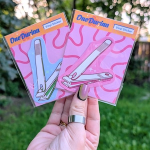 Peut inclure: Deux autocollants en vinyle représentant des coupe-ongles. L'un a un design bleu et blanc, l'autre un design rose et blanc. Les deux sont sur des fonds roses avec le texte "One Durian" et "Vinyl Sticker".
