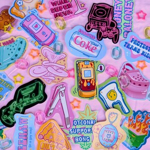 May include: A collection of colorful stickers and charms on a pink background. Stickers include a dog, a wine glass, a game console, a Diet Coke can, a cell phone, and a pizza slice. The text on the stickers includes "Red Wine Super Nova" and "Protect Trans Kids."