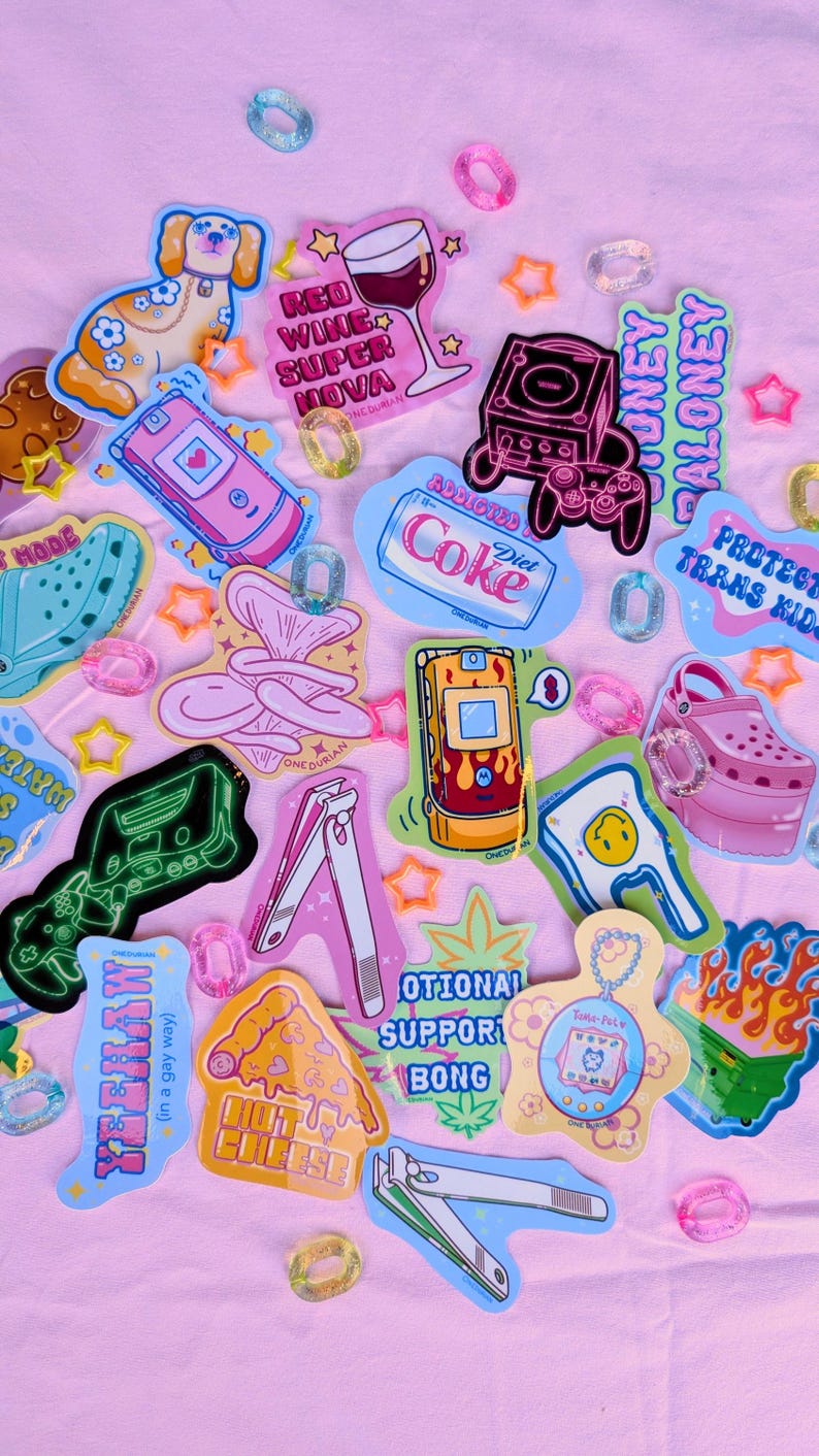 May include: A collection of colorful stickers and charms on a pink background. Stickers include a dog, a wine glass, a game console, a Diet Coke can, a cell phone, and a pizza slice. The text on the stickers includes "Red Wine Super Nova" and "Protect Trans Kids."