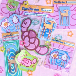 May include: A collection of colorful laminated magnetic bookmarks with whimsical illustrations. Designs include a worm, a dog, and a fried egg character. The bookmarks are packaged on pink cards with the brand name "One Durian".