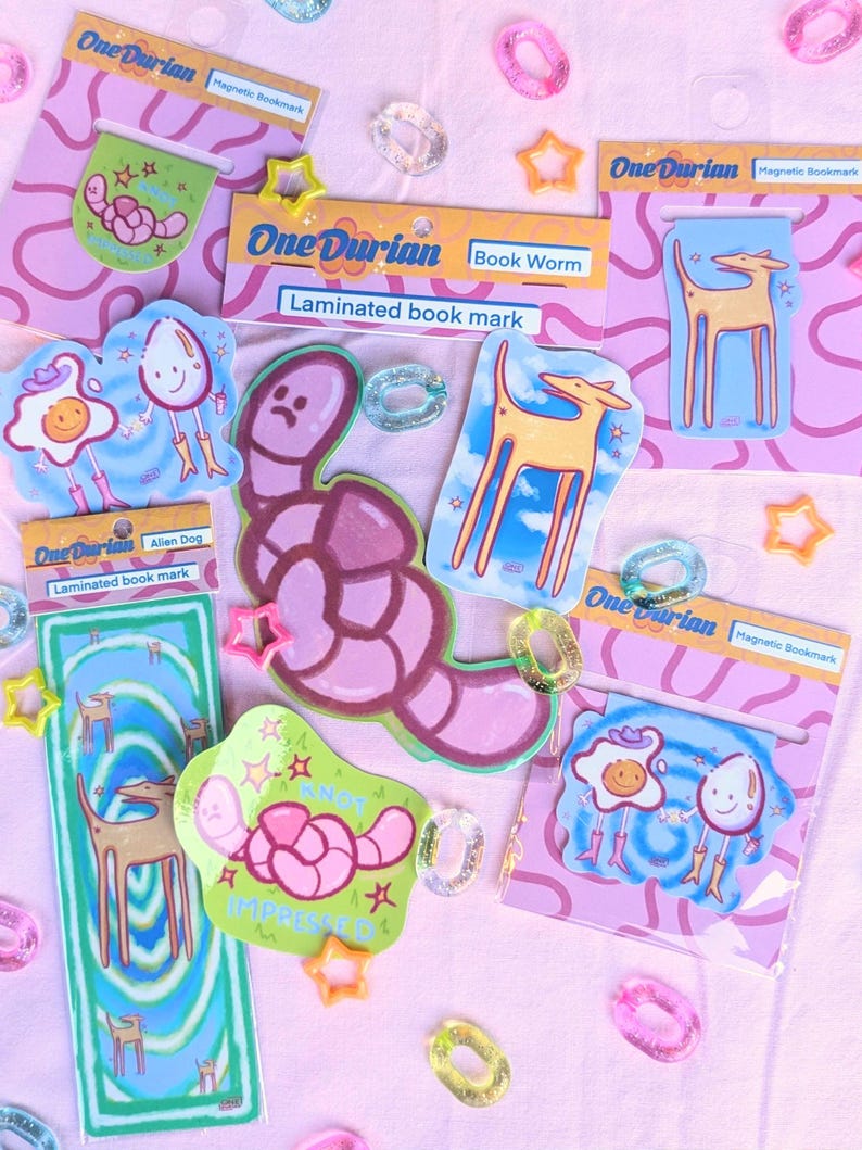 May include: A collection of colorful laminated magnetic bookmarks with whimsical illustrations. Designs include a worm, a dog, and a fried egg character. The bookmarks are packaged on pink cards with the brand name "One Durian".