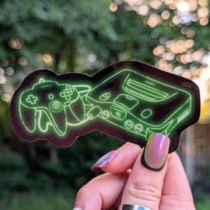 May include: A sticker with a neon green outline of a retro video game console and controller. The sticker has a black background and is held up against a blurred green background.