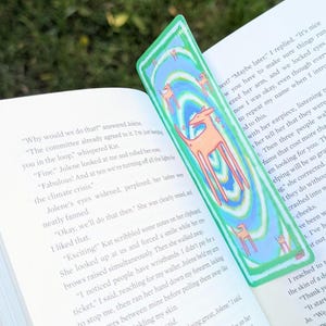 May include: A green and blue bookmark with a repeating pattern of orange deer and swirling lines. The bookmark is placed in an open book with text visible on the pages. The bookmark is a unique accessory for book lovers.