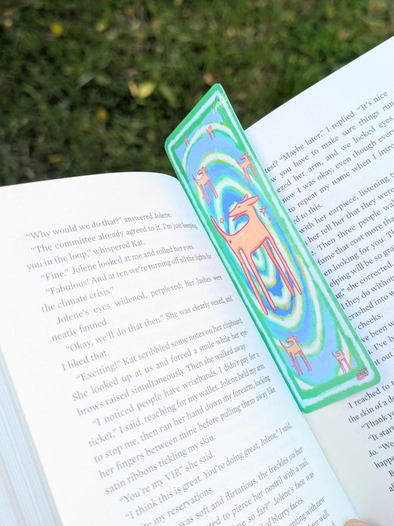 May include: A green and blue bookmark with a repeating pattern of orange deer and swirling lines. The bookmark is placed in an open book with text visible on the pages. The bookmark is a unique accessory for book lovers.