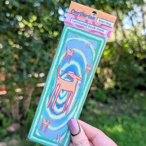 May include: Laminated bookmark featuring an alien dog design. The bookmark has a green border and a blue and green background with a spiral pattern. The text "One Durian" and "Alien Dog" are printed on the top.