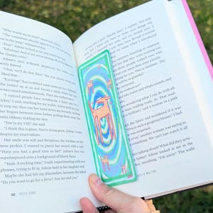 May include: A colorful bookmark featuring a deer design with a blue, green, and orange color scheme. The bookmark is rectangular and held over an open book with text visible.