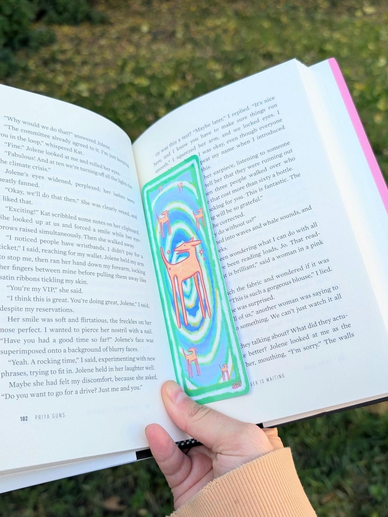 May include: A colorful bookmark featuring a deer design with a blue, green, and orange color scheme. The bookmark is rectangular and held over an open book with text visible.