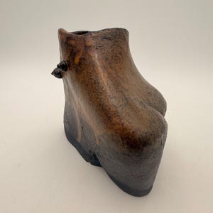 Puede incluir: Un jarrón de cerámica hecho a mano con forma de bota. El jarrón presenta una combinación de colores marrón oscuro y bronce con una superficie texturizada. Dos pequeños botones oscuros están en el lateral. La base es de color oscuro y sólido.