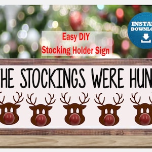 Puede incluir: Un letrero de madera blanco con el texto "The Stockings Were Hung" y cinco siluetas de renos con narices rojas. El letrero es un soporte para calcetines DIY.