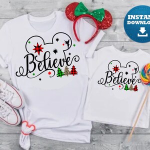 May include: Two white t-shirts with a black Mickey Mouse head outline and the word "Believe" in a black cursive font. The Mickey Mouse head has red and green stars and a small Tinkerbell silhouette. There are also small green and red plaid Christmas trees with a leopard print tree in the middle.