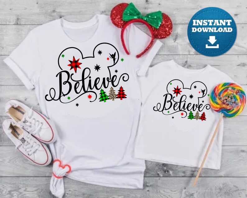May include: Two white t-shirts with a black Mickey Mouse head outline and the word "Believe" in a black cursive font. The Mickey Mouse head has red and green stars and a small Tinkerbell silhouette. There are also small green and red plaid Christmas trees with a leopard print tree in the middle.